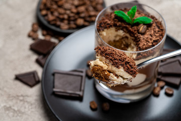 Classic tiramisu dessert in a glass cup, pieces of chocolate on concrete background