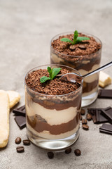 Classic tiramisu dessert in a glass cup, pieces of chocolate and savoiardi cookies on concrete background