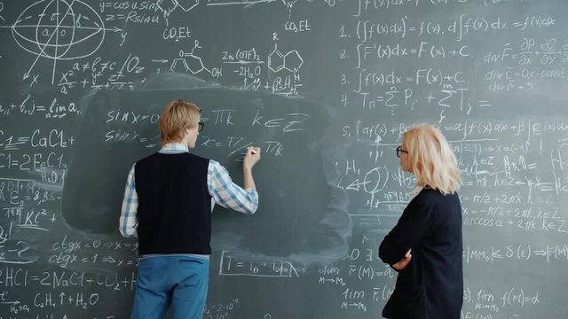 Student and helpful female teacher are working with chalkboard writing formulas in classroom, woman is teaching the guy talking explaining information.