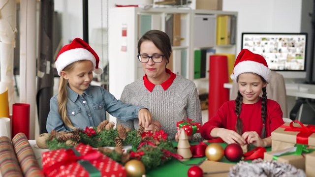 Woman and two little girls in Santa hats smiling and talking while preparing handmade Christmas gifts during workshop in creative studio