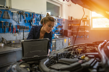 An attractive woman mechanic working on a car in a repair shop. A female mechanic is working under a bonnet of a car in a garage repair shop. She is wearing blue overalls.