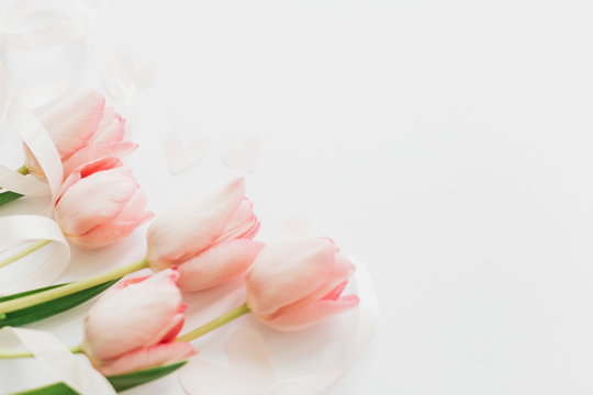 Happy Mothers day. Pink tulips with ribbon and hearts on white background. Stylish soft spring image. Happy womens day. Greeting card mockup with space for text.  Hello spring