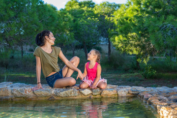 Beautiful mother and her little daughter outdoors. Beauty Mum and her Child playing with water in Park together at sunset. Outdoor Portrait of happy family. Mother's Day