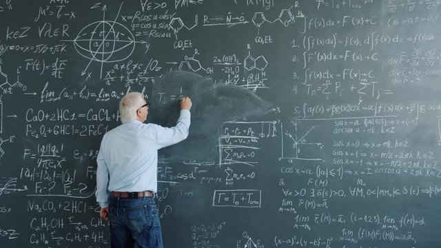 Mature man physicist writing formulas on blackboard with chalk indoors at school busy with solving scientific problem. Science, occupation and education concept.