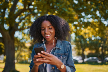 Portrait of smiling young african american woman listening to music on earphone using mobile phone in the park - Happy young black woman listening to music in the park on a sunny day 