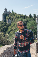 Man dressed in a motorcycle outfit and sunglasses, sport watch. body protection turtle, helmet in hand. Fortress, stone stairs on background. Vertical photo. Pass of the witches San Marino