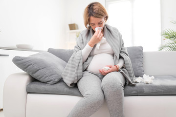 Pregnant woman catching cold, flu, virus, sitting at home on a couch, blowing nose and being sick.