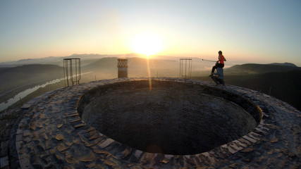 Couple on top of high smoke stack chimney doing acrobatics with the risk of falling thousand of meters to the ground, golden sunrise in the horizon