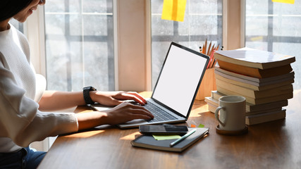 Cropped image of young beautiful woman working as writer typing on computer laptop with white blank screen while sitting at the wooden working table with sunlight through windows as background. - obrazy, fototapety, plakaty
