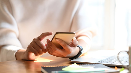 Cropped image of creative woman working as graphic designer holding on smartphone while sitting at the modern working table with comfortable living room as background.