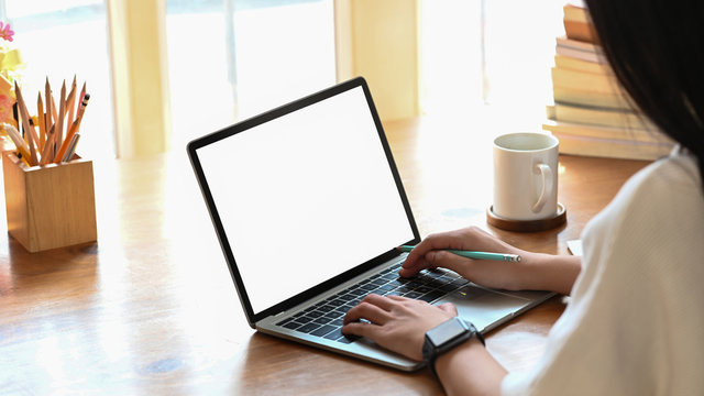Cropped image of young beautiful woman working as writer typing on computer laptop with white blank screen while sitting at the wooden working table with sunlight through windows as background.