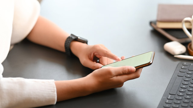 Cropped image of creative woman working as graphic designer holding on smartphone while sitting at the modern working table with comfortable living room as background.