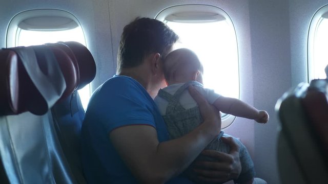 Man hold little baby against airliner window, try to entertain tiny passenger when mother go away for a while. Young family travel by economy class of regular airline.