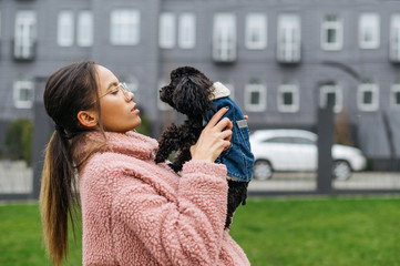 Portrait of attractive girl with beautiful dog breed toy poodle in hands on street landscape background, woman wearing pink jacket. Beautiful lady spends her free time with her pet outdoors.