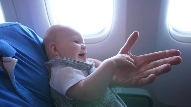 Little baby sit on hands of father during airline flight, play with large arm of dad. Tiny passenger girl enjoy easy travel time, flap small hand on man palm