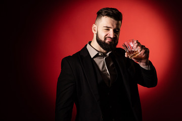 Fototapeta na wymiar Handsome bearded man in suit with stylish beard holding a glass of whiskey and going to drink it, on red background