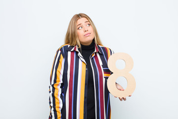 young pretty blonde woman confused, doubtful, thinking, holding a number 8.