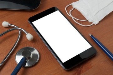 Blank screen smartphone surrounded by a stethoscope, a medical mask and a pen on a wooden table