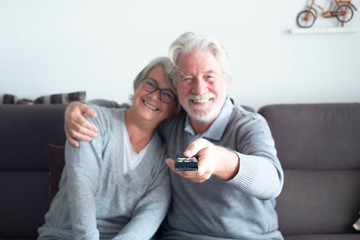 couple of pensioners seniors looking and watching the TV together at home sitting on the sofa - two mature people smiling and man changing channel with the remote control