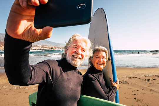 couple of pensioners seniors taking a selfie together at the beach with their wetsuits and surfboards - mature people learning surfing