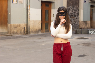 Fototapeta premium Portrait of attractive caucasian young woman with mask holding index finger near his lips, in the city, white top and red pants, dark air. Place for your text in copy space.