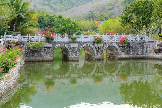 Stone bridge over a green pond surrounded by temples, gazebos, mountains, fowers, palm grove and stone fence on the territory of Buddhist center Nanshan.