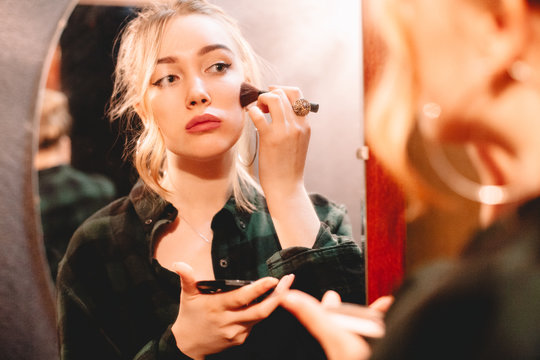 Young woman contouring her face applying make up with makeup brush looking at herself in the mirror at home getting ready to go out