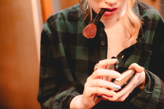 Young Woman Opening Palette And Holding Makeup Brush In Her Mouth While Applying Make Up At Home Getting Ready To Go Out