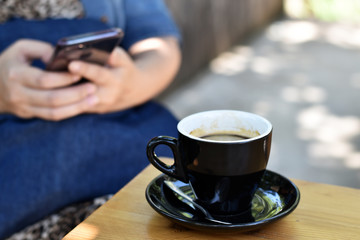 Black coffee cup on wooden table, Blurred woman sitting on a mobile.