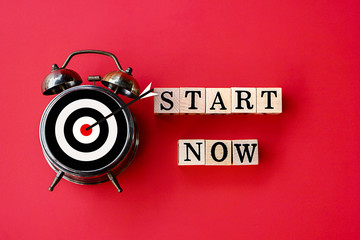 start now concept with text on wooden block and alarm clock on red background 