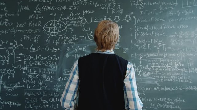 Portrait of intelligent guy in casual clothing walking to chalkboard writing formulas teaching science. Profession, lifestyle and knowledge concept.