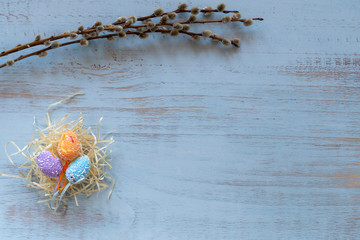 Colorful easter eggs in hay nest and willow branches laying on a blue wooden background. Easter holidays concept. Copyspace, place for text and wording. April vibes.