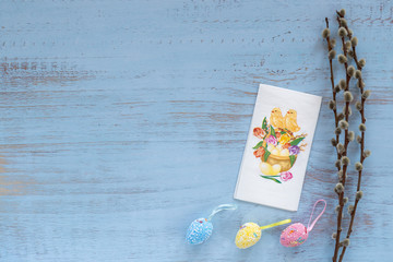 Napkin with a chicken, colorful easter eggs and willow branches laying isolated on a blue wooden background. Easter holidays concept. Copyspace, place for text and wording. April vibes.
