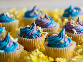 Yellow cupcakes with blue and purple frosting and confetti sprinkles sitting on a bed of yellow paper shreds with a white background, for a birthday or celebration, delicious and colorful.