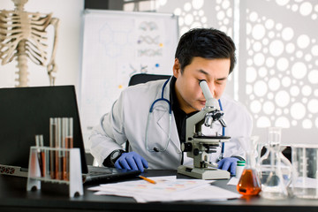 Shot of a Korean young man scientist looking through the lens of a microscope while sitting at the table with laptop, test tubes and flasks. Science, biology, medicine concept