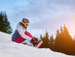 Horizontal snapshot of a young girl freerider, sitting on a slope in the mountains putting her...