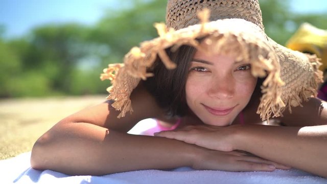SEAMLESS LOOP VIDEO: Asian woman lying down tanning wearing straw hat as sun protection on beach holiday. Summer travel beautiful young biracial girl relaxing smiling.