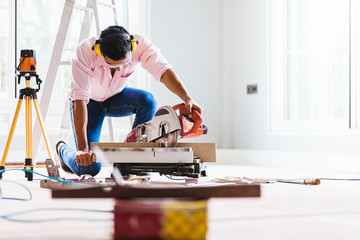 Obraz premium Smart indian contractor hand using sawing machine wood work and working overall equals a wooden bar in the house renovation background. Home improvement ideas concept.