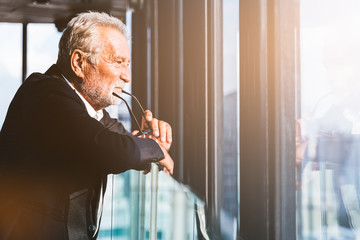 Retirement concept. Senior grey-haired businessman standing and looking to right hand at modern business lounge high up in an office tower.