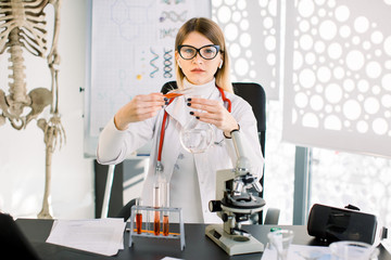 Pretty Caucasian woman researcher doctor chemist with glass equipment in the lab. Female professional scientist purifying red solution form tube into glass flask. Laboratory, clinical analysis concept