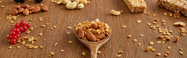 Wooden spatula with granola next to redcurrants, nuts and cereal bars on wooden background, panoramic shot