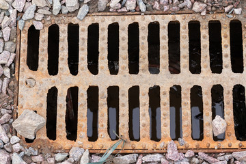 Rusty sewer grill with water beneath it, located on gravel covered ground