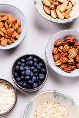 Ingredients for homemade granola: oatmeal, coconut crunch, pecans, almond, brazilan nuts, blueberries in a bowls on light background. Healthy diet breakfast. Flatlay. Top view. Selective focus
