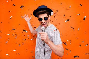 Photo of attractive crazy funky youngster guy singing karaoke using microphone confetti falling wear retro hat sun specs striped t-shirt isolated bright orange color background