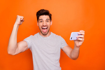 Photo of cool excited guy good mood hold telephone raise fist celebrate first startup successful income wear striped t-shirt isolated bright orange color background