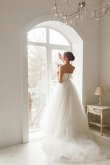 Fototapeta premium Young bride in a beautiful dress holding a bouquet of flowers posing near window in bright white studio. Wedding concept.