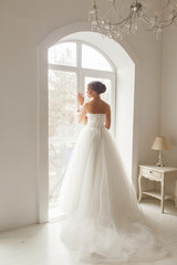 Young bride in a beautiful dress holding a bouquet of flowers posing near window in bright white studio. Wedding concept.