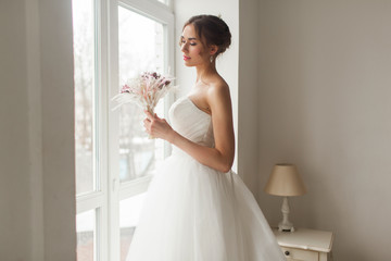 Young bride in a beautiful dress holding a bouquet of flowers posing near window in bright white studio. Wedding concept.