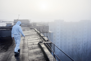 Back view of man wearing white coverall and gas mask, walking on the roof of an urban building. On...