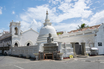 A modest buddhist white temple with a buddha statue in the foreground and against a blue sky with...